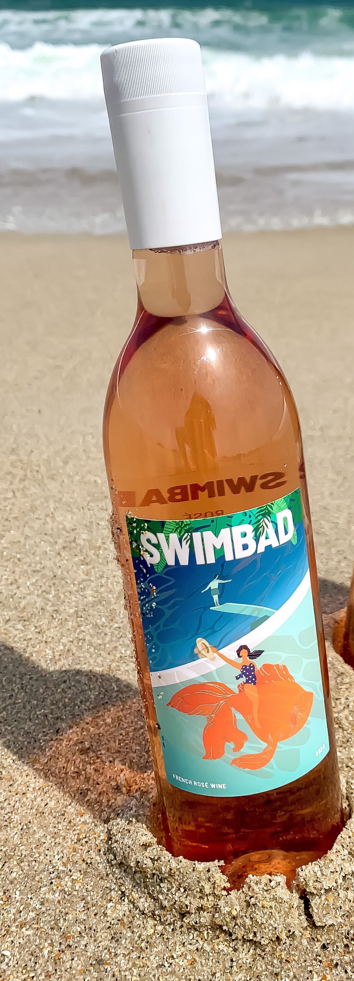 A Swimbad rosé PET bottle standing in beach sand with surf behind it